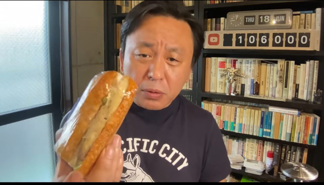 本棚を背景に、タレントの菅野完がラップに包まれたサンドイッチを手に持ち、カメラに見せている。