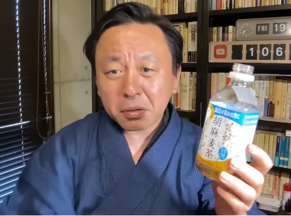 青い着物を着た菅野完が、手元に胡麻麦茶のペットボトルを持ち、真剣な表情でカメラを見つめている様子。