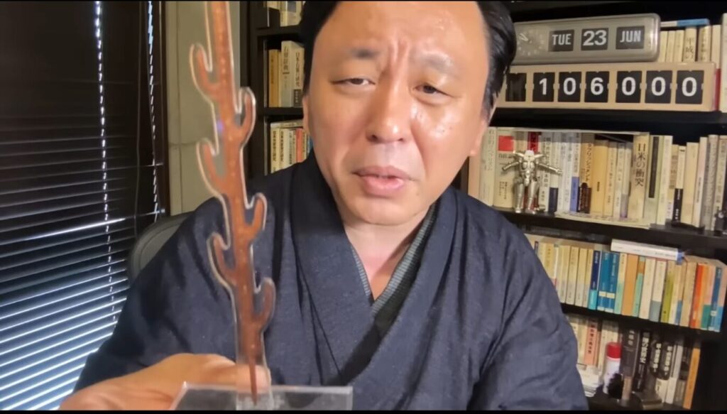 着物を着た菅野完が、手前で珊瑚のような形をした透明なアクリルスタンドを掲げながら、背景の書棚を背にして語りかけている様子。