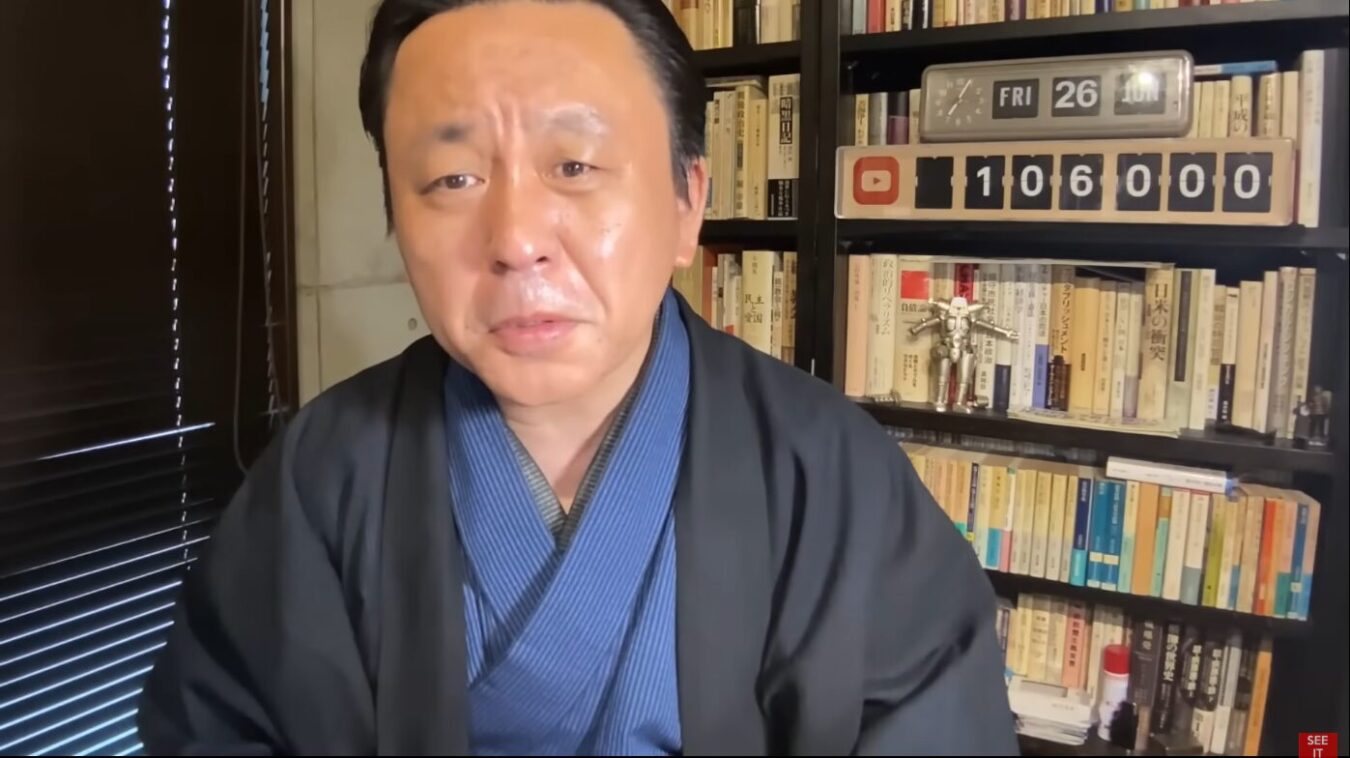 和服姿の菅野完が本棚を背景に真剣な表情でカメラを見据えている様子。背後には大量の本と登録者数を示すカウンターが置かれている。