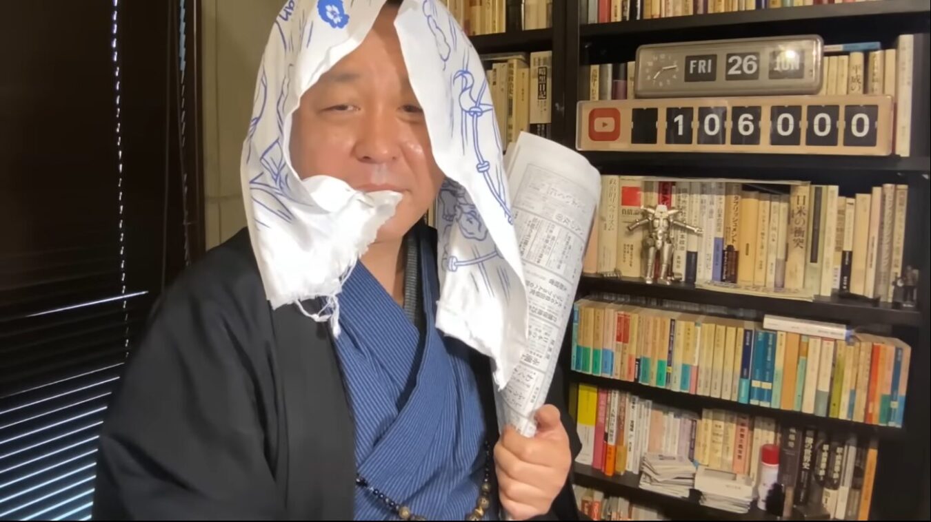 和服を着て手ぬぐいを頭にかぶり、新聞紙と紙くわえをした男性が、本棚の前でポーズをとっている様子。
