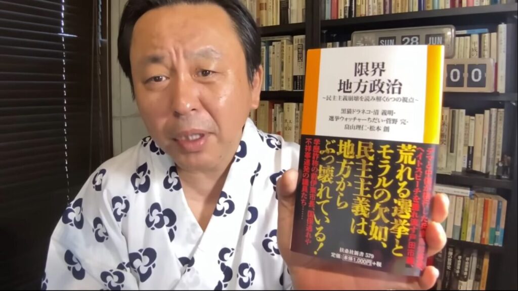 本棚を背景に、浴衣姿の菅野完が新書『限界地方政治』を手に持ち、カメラに向かって語りかけている様子。