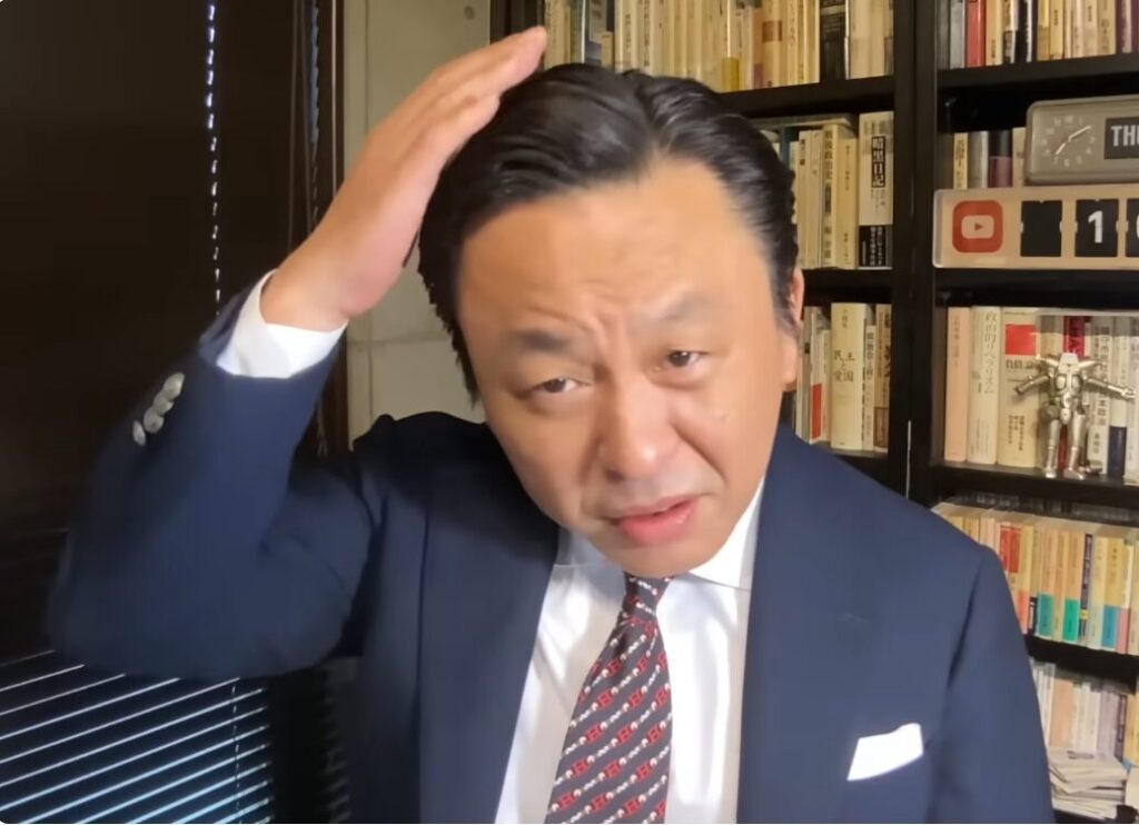 本棚が並ぶ書斎を背景に、スーツ姿の菅野完が右手を頭に添え、カメラを見つめる様子