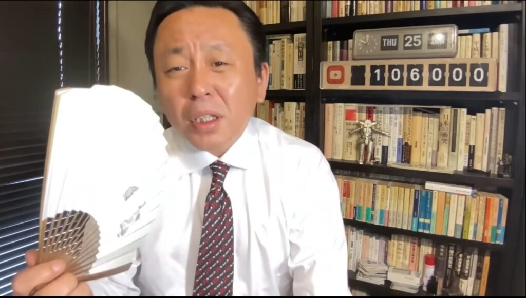 書斎の本棚を背景に、白いワイシャツとネクタイを着た菅野完が、扇子を手に持ち熱心に語りかける様子。