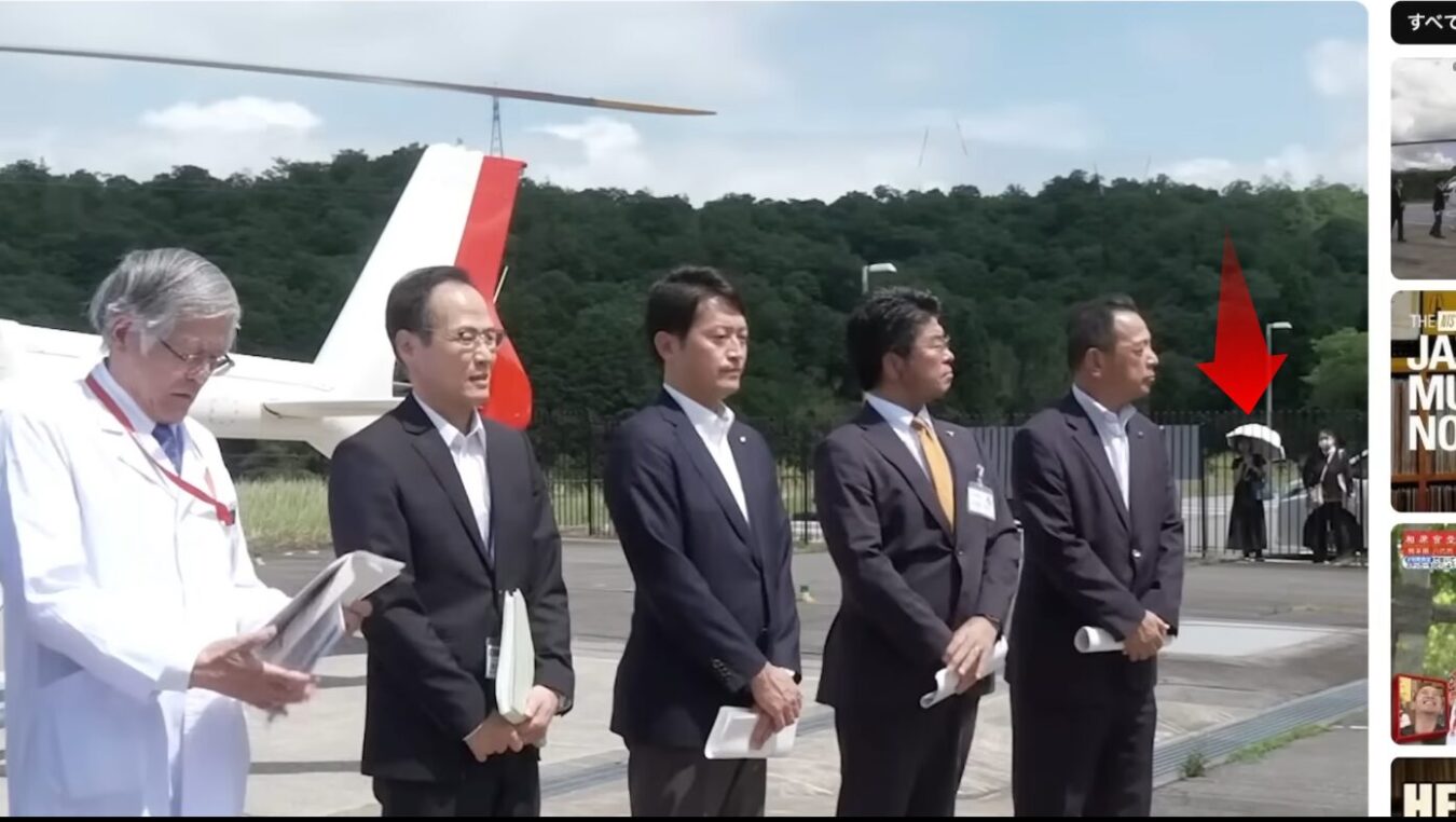 ヘリコプターの前に並ぶ白衣とスーツ姿の男性たち。背景にいる傘を持った人物が赤い矢印で指されている。