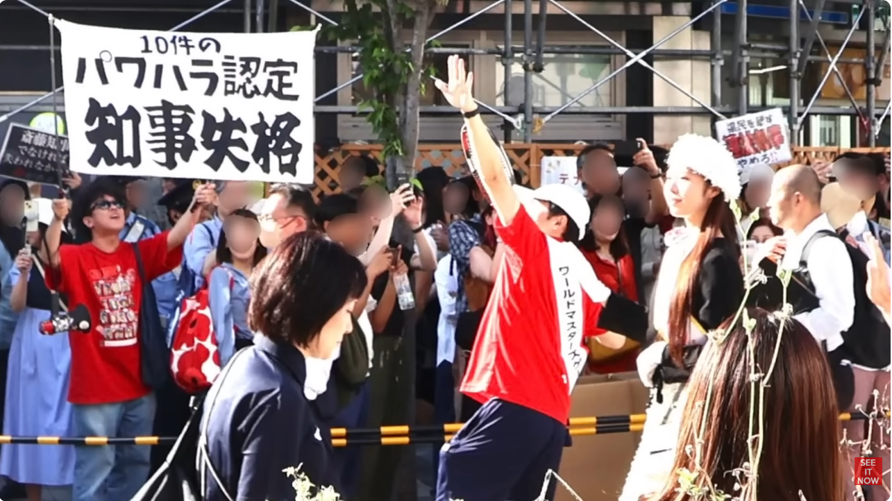 路上で「パワハラ認定 知事失格」と書かれた横断幕を掲げて抗議活動を行う人々と群衆。