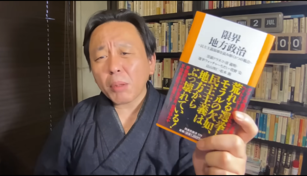 和服姿の菅野完が、地方政治の現状を批判する内容が書かれた書籍を手に持ち、カメラに向かって語りかけている様子。