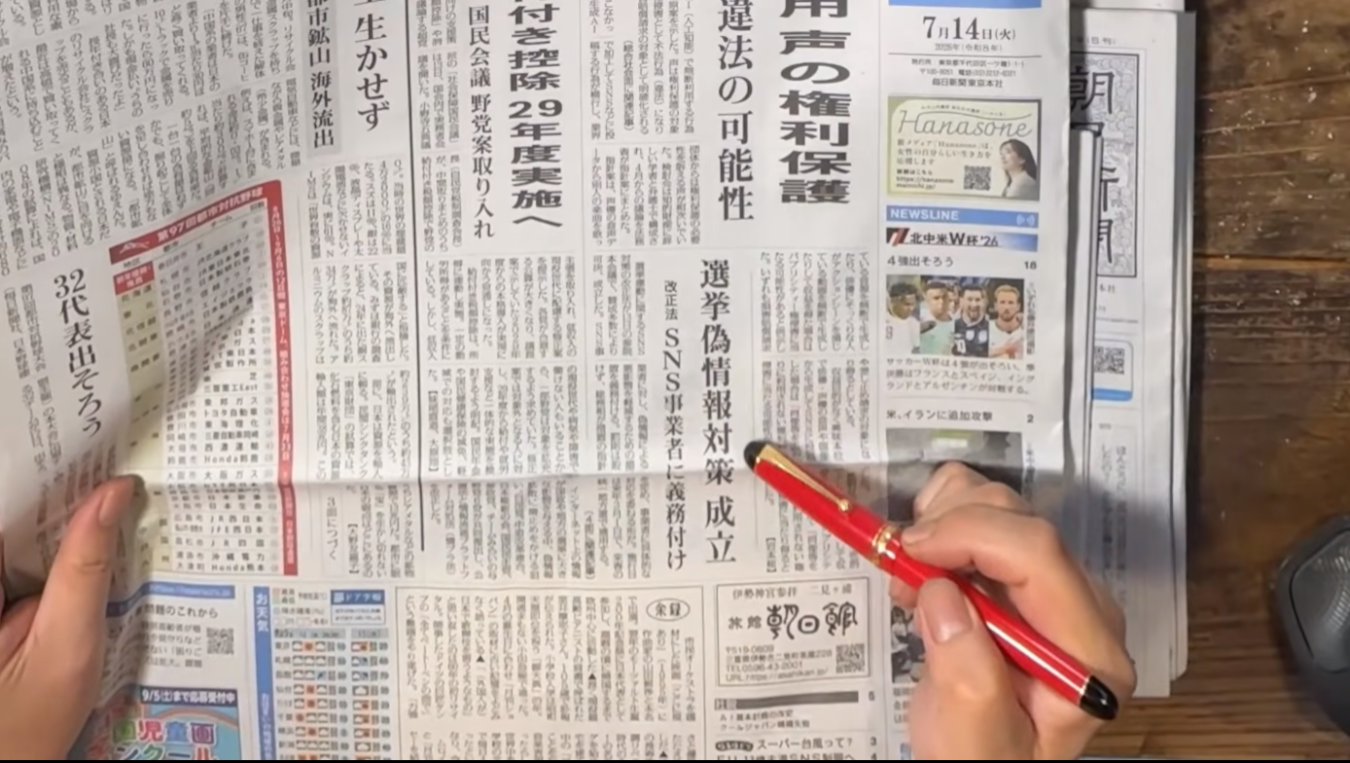 新聞の記事の見出し「選挙偽情報対策成立」を赤いペンの先で指し示している手。