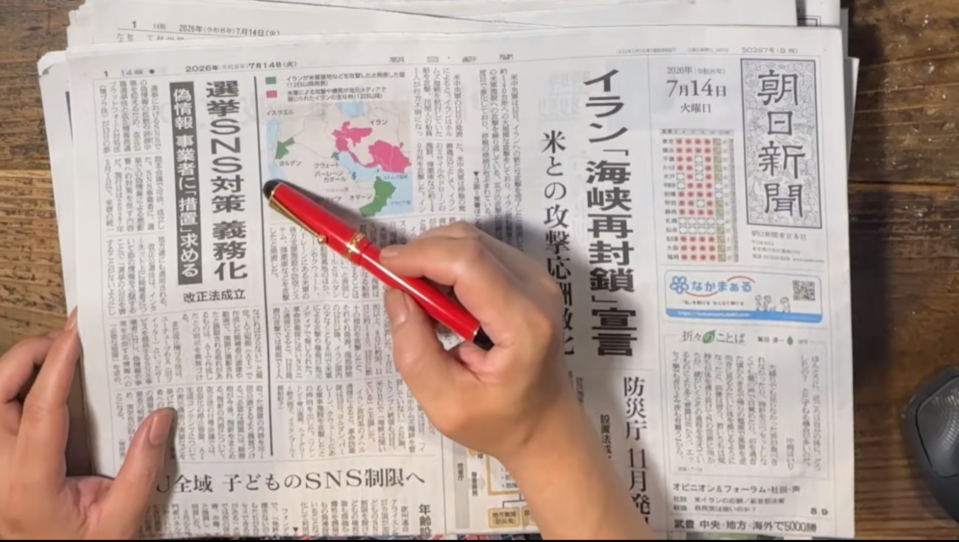 赤いペンを持つ手で日本の新聞紙面の一面を指し示している様子