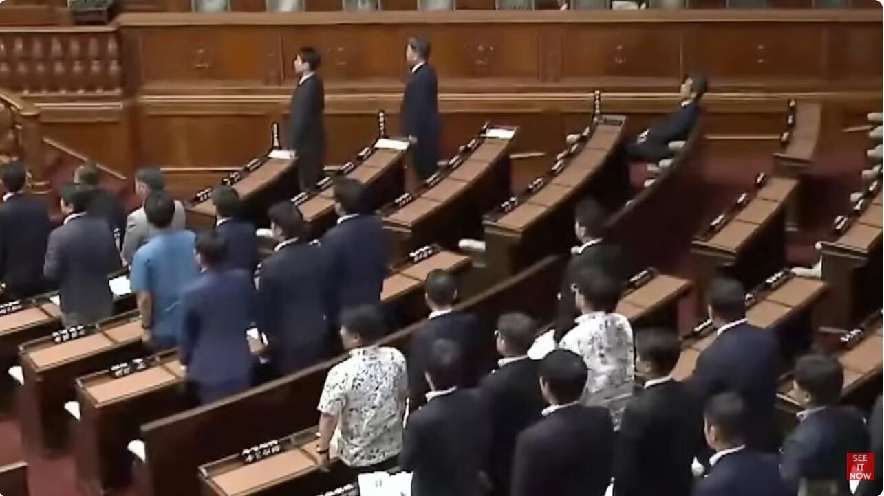 国会議事堂のような議場で、スーツ姿の議員たちが整列して起立している様子。