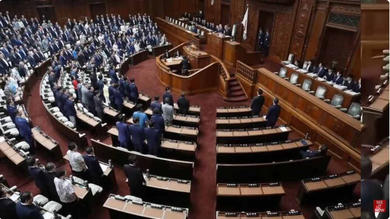 多くの議員が着席または起立している、重厚な木造建築の日本の国会議事堂の議場内風景。