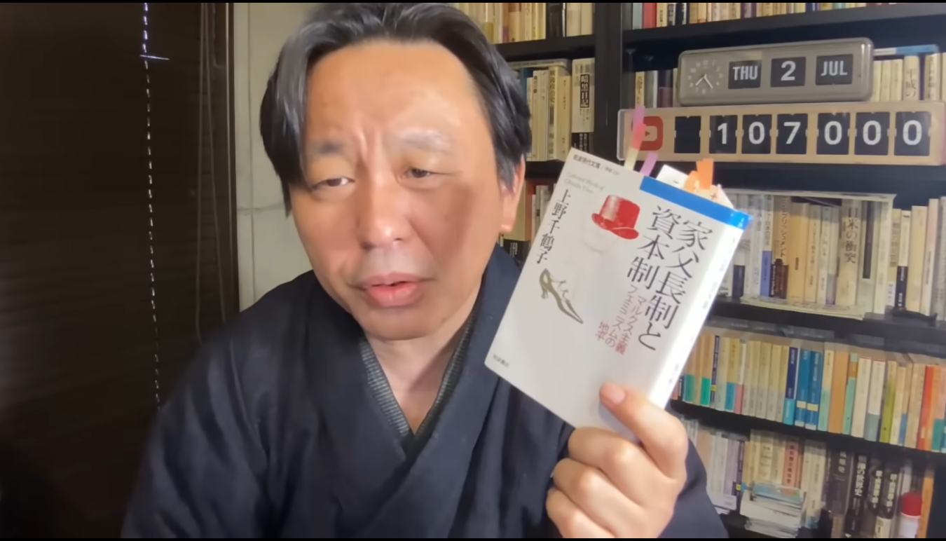 和服姿の菅野完が、上野千鶴子著『家父長制と資本制』というタイトルの本を手に持ち、カメラに向けて紹介している様子。