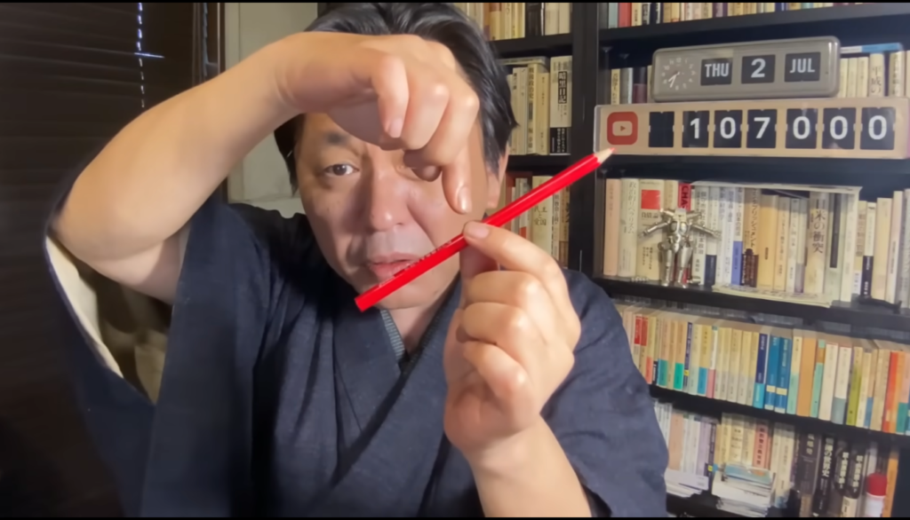 着物を着た男性が、赤いペンを指で持ちながらカメラに向かって何かを説明している様子。