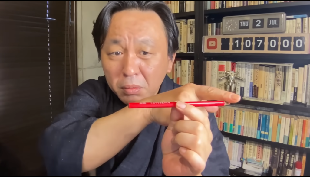 書斎で赤い鉛筆を指先に乗せてバランスをとる、和服姿の男性の様子。