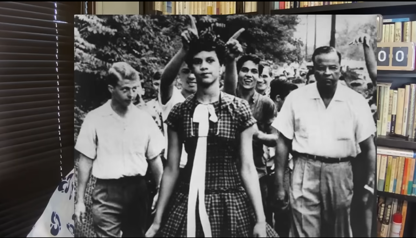 1950年代、人種差別に抗議し学校へ向かう黒人女子生徒と、彼女を先導する男性や周囲の学生たちの白黒写真。