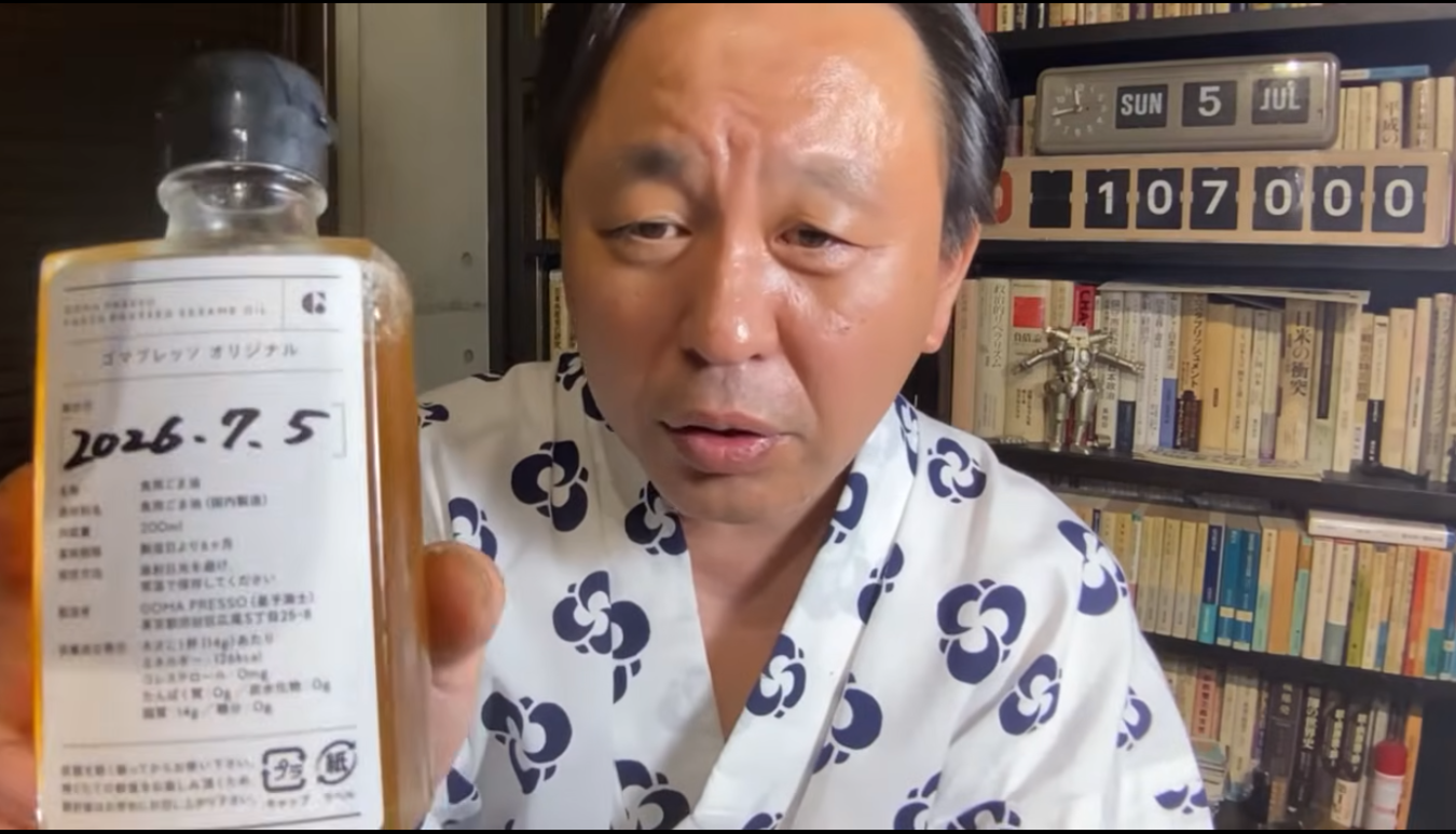 浴衣姿の菅野完が、賞味期限が2026年7月5日と記載されたゴマプレッソの純正ごま油のボトルを手に持っている様子。