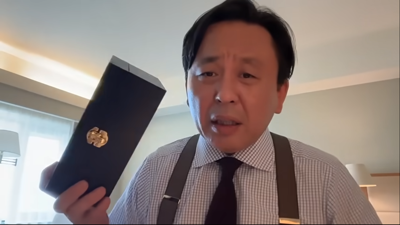 チェック柄のシャツとサスペンダーを着用した菅野完が、金色の留め具が付いた黒い長方形の箱を手に持って話している様子。