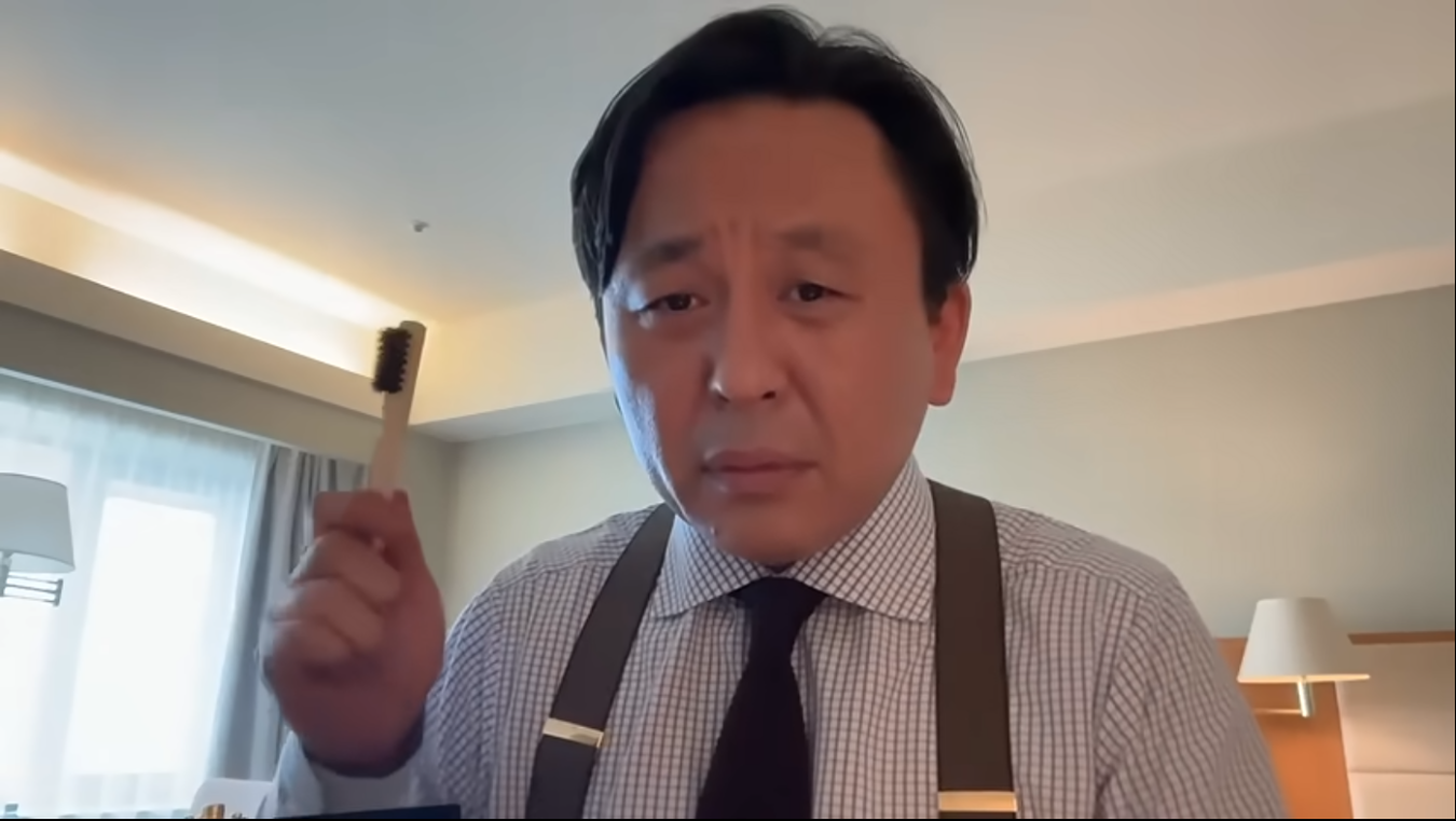 チェック柄のシャツとサスペンダーを着用し、手元に歯ブラシを持ってカメラを見つめる菅野完。