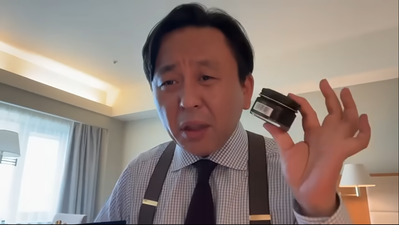 チェック柄のシャツとサスペンダーを着用した菅野完が、黒い蓋の小さな容器を手に持って説明している様子。