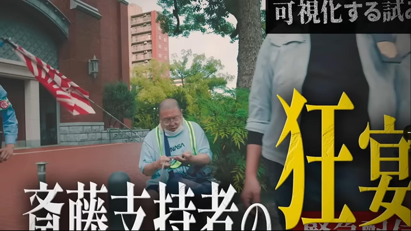 街頭でNASAのTシャツを着た男性が作業し、その手前に「斎藤支持者の狂宴」という大きな文字が重なる様子。