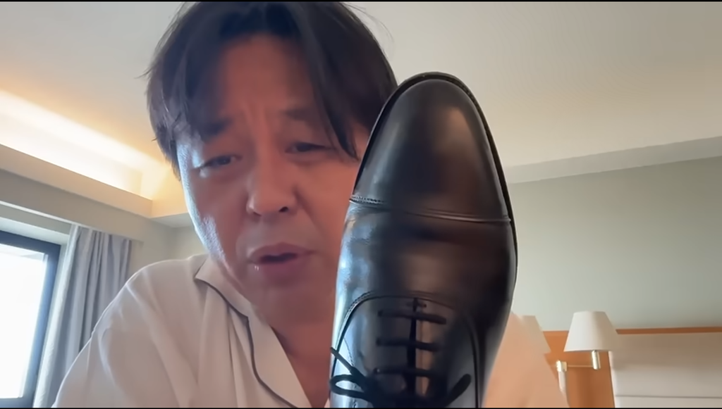 ホテルの部屋で、カメラに向かって黒い革靴のつま先を見せながら話す菅野完。