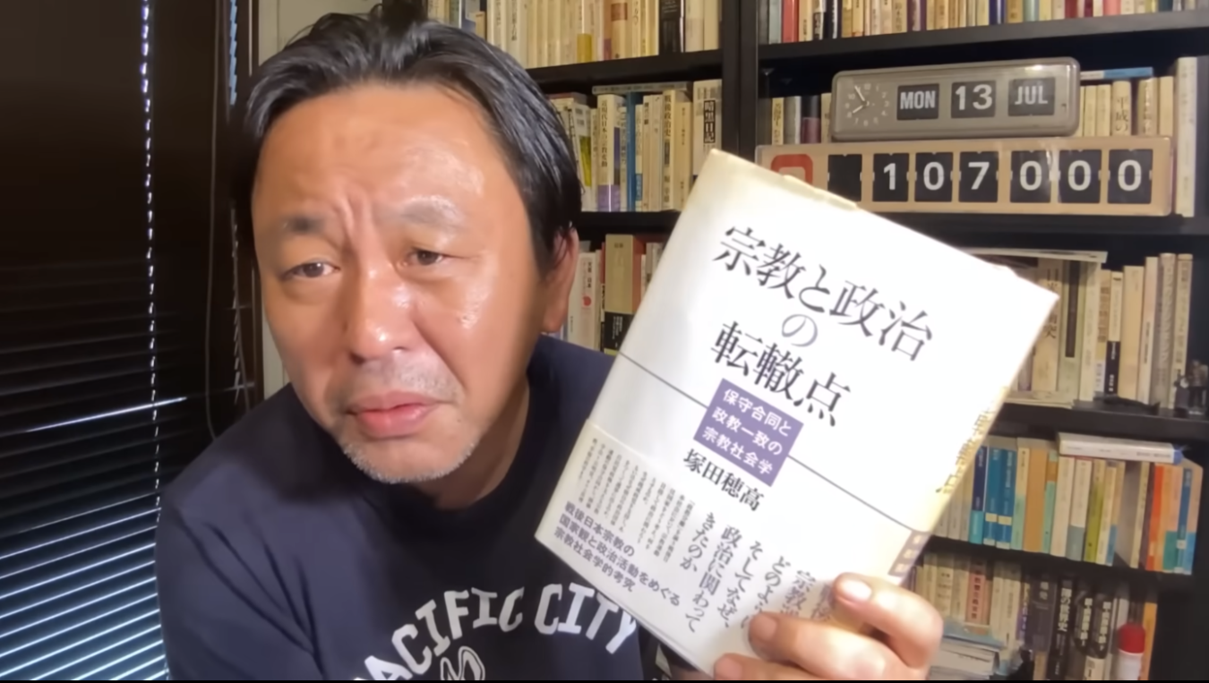 菅野完が本を持ちながらカメラを見つめている様子